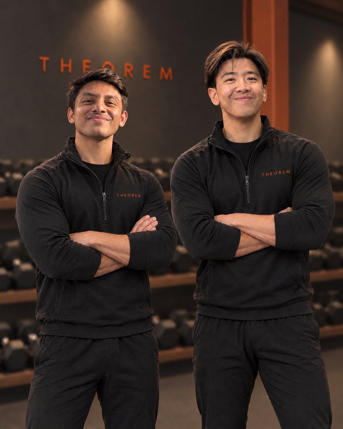 Kulan and Kevin, the founders, arms folded in black Theorem quarter-zips in front of the dumbbell rack under the Theorem sign
